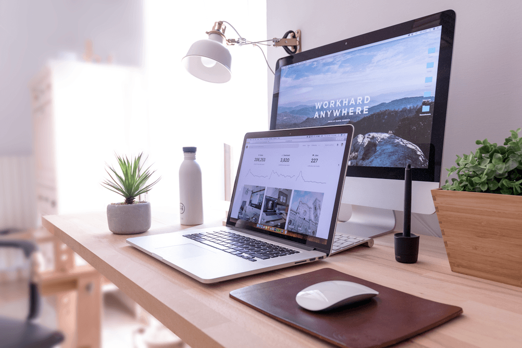 Bureau de travail minimaliste avec ordinateur portable, écran externe, souris et plante sur un bureau en bois lumineux.